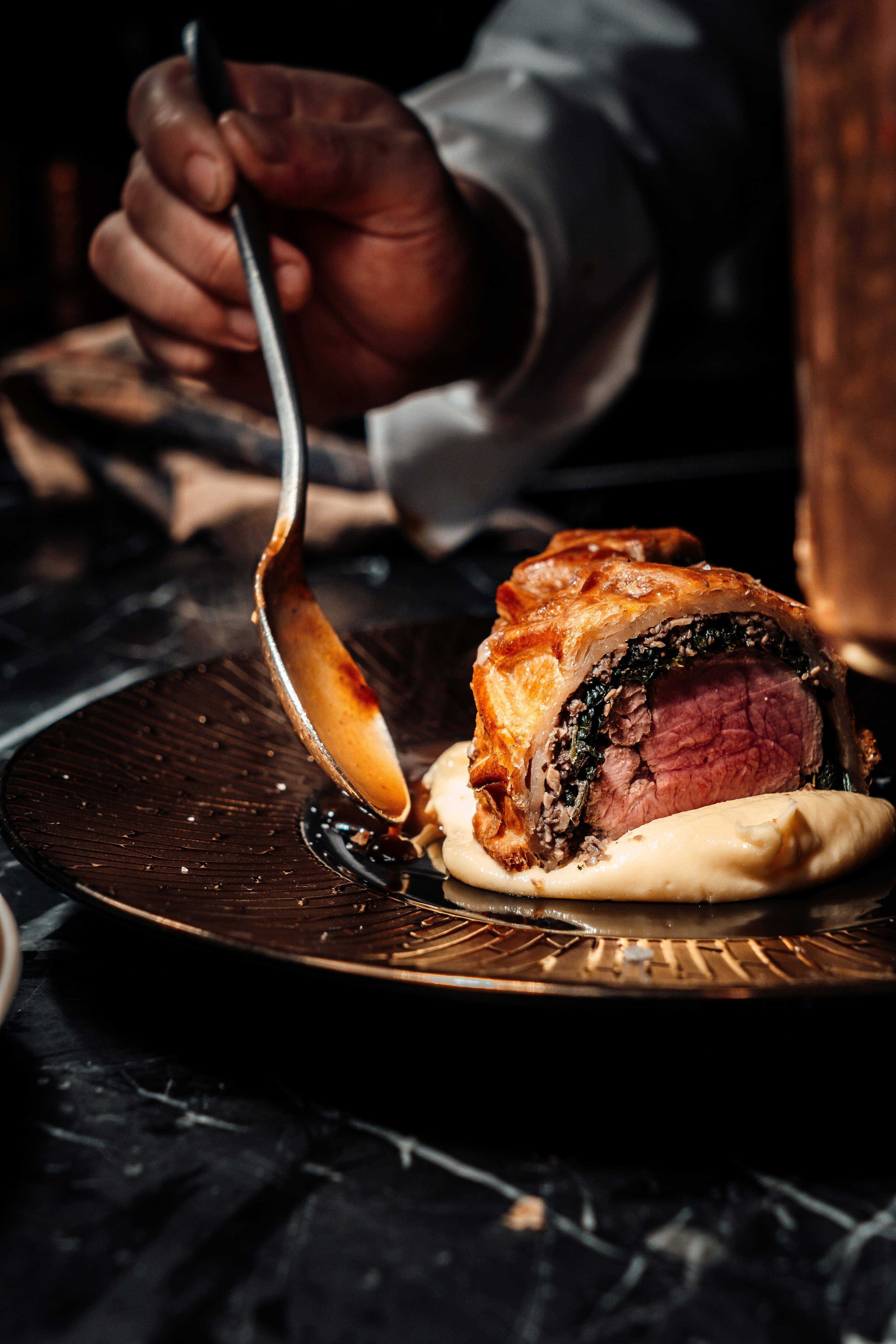 A chef ladling sauce over a sliced beef wellington on a dark plate